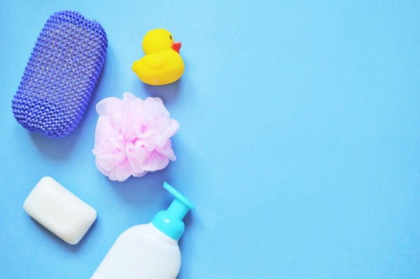 A collection of bath items including a yellow rubber duck, purple sponge, pink bath pouf, bar of soap, and a bottle of liquid soap against a blue background.