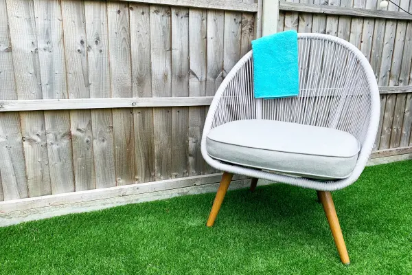 A white modern chair with a teal towel on the armrest, on artificial grass against a wooden fence.