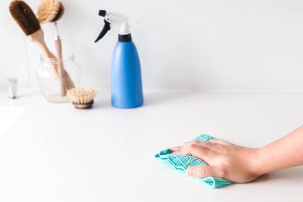 A hand wiping a surface with a cloth, a spray bottle, and cleaning brushes in the background.