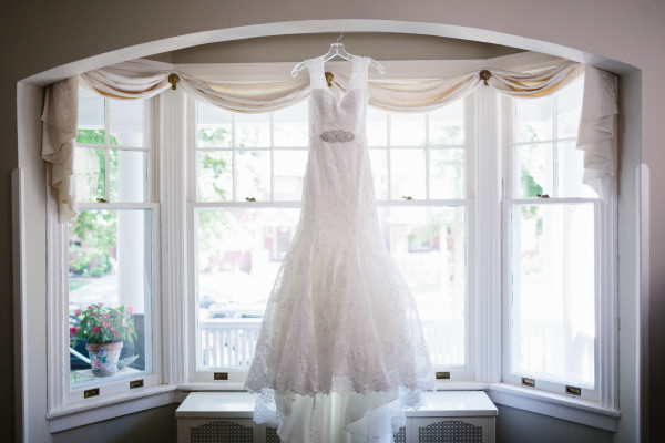 A wedding dress hanging in front of a window with sheer curtains and a flower pot on the windowsill.