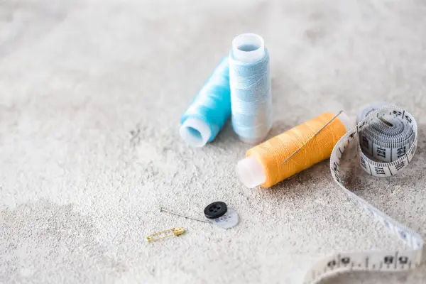 Sewing supplies including thread spools, a needle, a button, a safety pin, and a measuring tape on a textured surface.