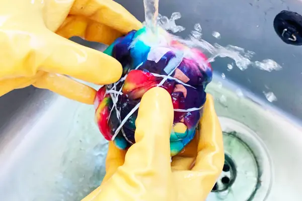 Hands in yellow rubber gloves rinsing a colourful fabric under running tap water in a sink.