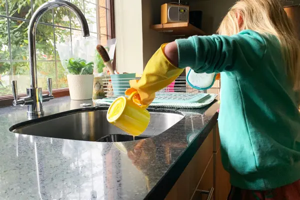 Person in yellow gloves pouring something into a kitchen sink.