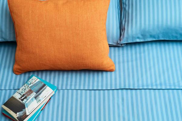 A neatly made bed with striped blue and white bedding, two pillows, and an orange cushion with a small stack of books beside it.