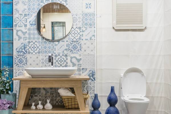 A tidy and clean bathroom with ornate blue and white tiles, a wooden vanity unit with a white basin, round mirror, toilet, and decorative blue vases.