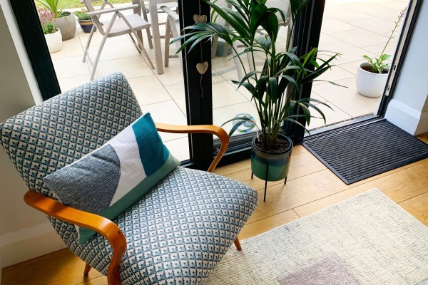 A tidy room with a patterned armchair, decorative cushion, potted plant, and a clean rug near an open door leading to an outdoor seating area.