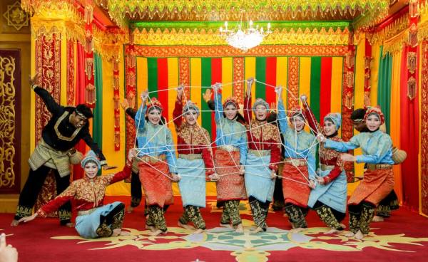 Sebuah kelompok penari mengenakan kostum tradisional melakukan pertunjukan budaya, dengan latar belakang dekorasi serba warna-warni dan emas.