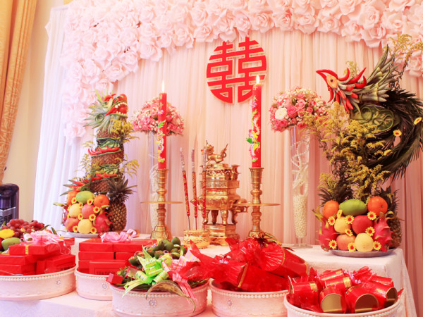 Đây là bức ảnh về bàn thờ cúng với trái cây, bánh kẹo, hương, đèn cầy và hoa, có sự trang trí theo phong cách Á Đông.