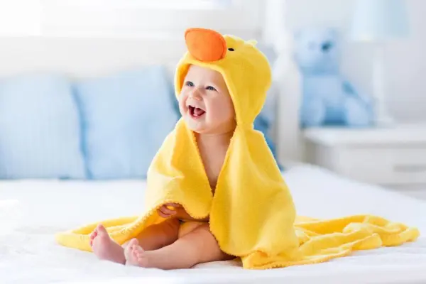 Em bé mặc khăn tắm màu vàng, ngồi cười trên giường.