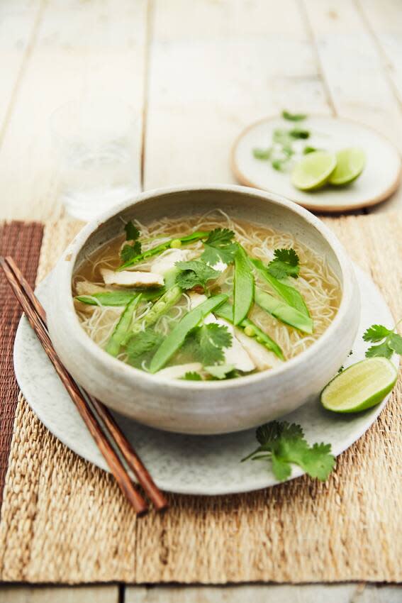 Một tô phở gà với rau mùi và đậu Hà Lan, cùng với một đĩa chanh và đũa bên cạnh trên bàn gỗ.