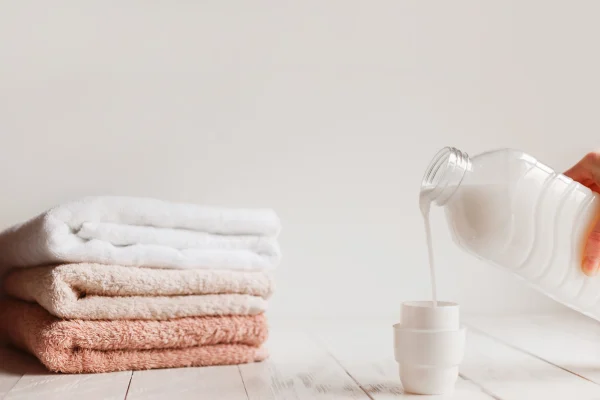 Một người đang đổ nước giặt vào nắp đong, cạnh chồng khăn tắm gấp ngăn nắp.