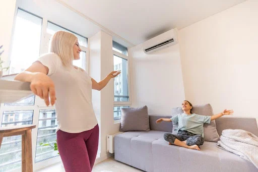 Phòng khách sạch sẽ với hai phụ nữ, một người đứng và một người ngồi trên ghế sofa, cùng quạt máy lạnh trên tường.