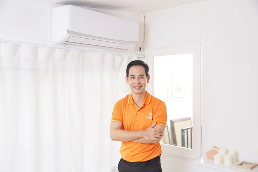 Người đàn ông mặc áo thun màu cam đứng tựa tay dưới máy điều hòa trong phòng.