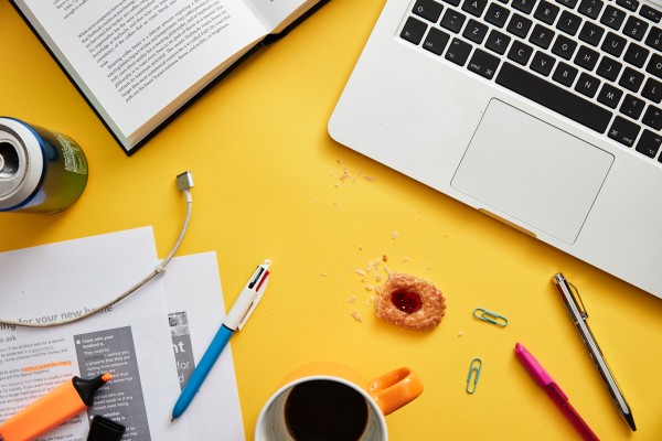 A cluttered desk with a laptop, coffee cup, spilled drink, papers, a pen, a USB cable, and a pastry with a bite taken out on a yellow surface.
