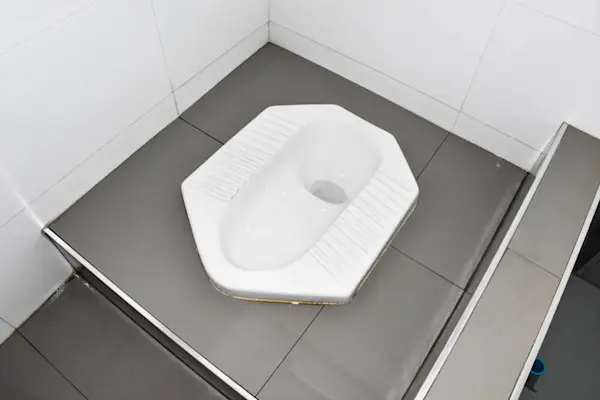 Toilet jongkok putih di kamar mandi dengan ubin abu-abu.