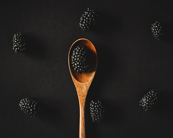 Ein Holzlöffel mit einer Brombeere auf schwarzem Hintergrund, umgeben von weiteren Brombeeren.