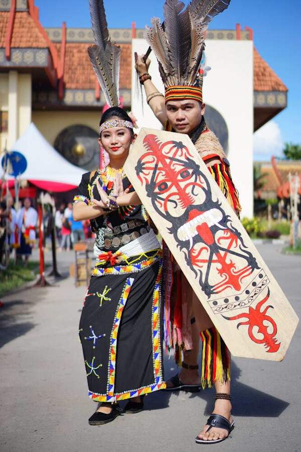 Dua orang mengenakan pakaian adat dan berpose dengan perisai tradisional di luar gedung.