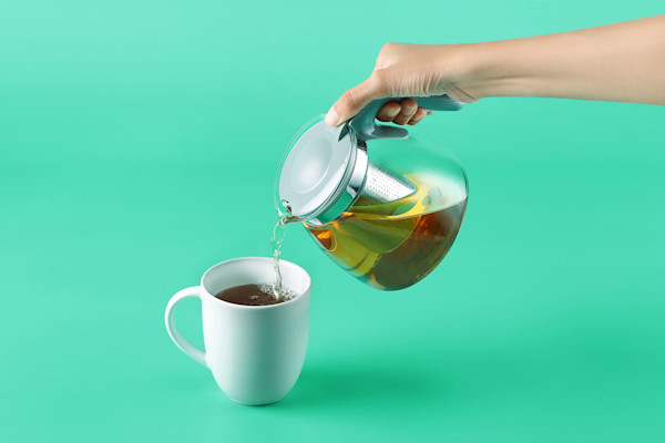 Eine Hand gießt Tee aus einer Teekanne in eine weiße Tasse vor einem türkisfarbenen Hintergrund.
