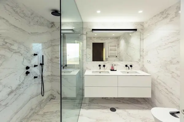 Kamar mandi modern dengan dinding dan lantai marmer, dilengkapi dengan shower, wastafel ganda, dan cermin.