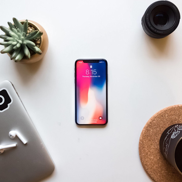 Auf dem Bild sind ein Smartphone, ein Laptop mit Aufkleber, ein Kameraobjektiv, eine sukulentenhaltende Topfpflanze und eine Untersetzer auf einem weißen Hintergrund zu sehen.