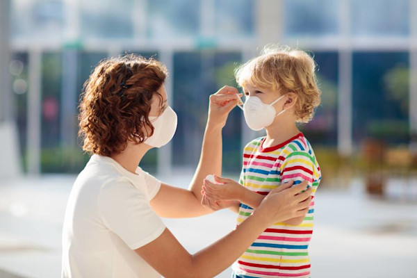 Seorang ibu sedang memakaikan masker pada anaknya di luar ruangan.