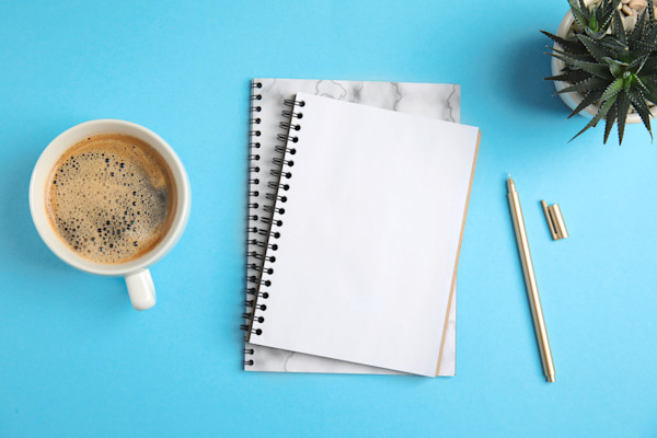 Een kopje koffie, een notitieblok met een pen en een vetplant op een blauwe achtergrond.