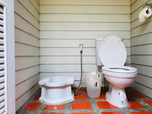 Kamar mandi luar ruangan dengan toilet dan bak cebok, serta ember dan tisu toilet.