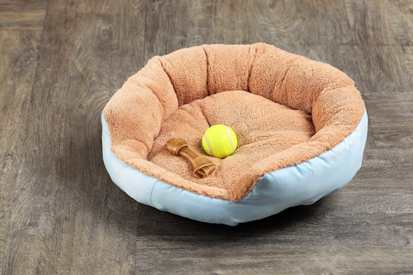 Una cama para mascotas con una pelota de tenis y dos huesos de juguete sobre un suelo de parquet.