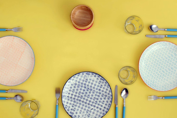 A flat lay of a table setting with colorful plates, glasses, and cutlery on a yellow background.