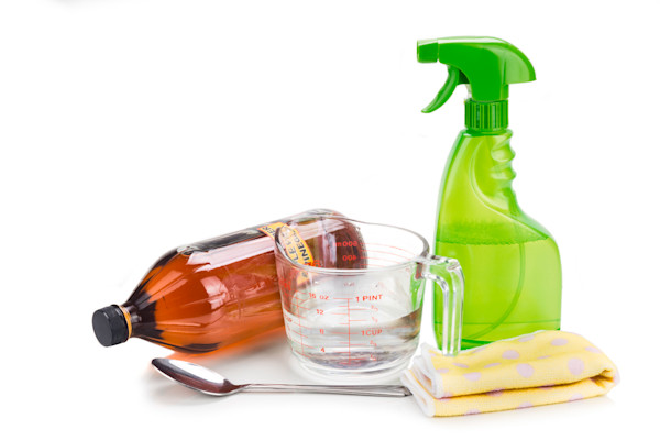 Cleaning supplies including a green spray bottle, a measuring cup, a brown bottle, a spoon, and a yellow cloth with white dots, all against a white background.