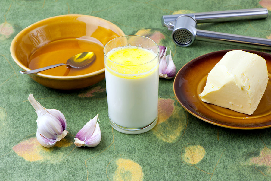 Ein leeres Geschirr mit Löffel, ein Glas Milch, ein Stück Butter, Knoblauchzehen und ein Knoblauchpresse auf einer gemusterten Tischdecke.