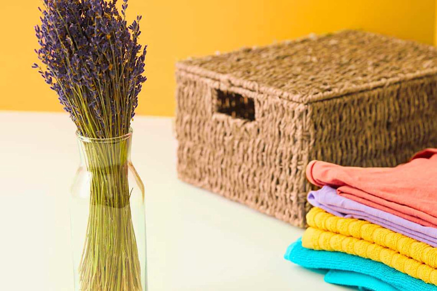 Ein Strauß Lavendel in einer Vase, eine geflochtene Aufbewahrungsbox und gestapelte Reinigungstücher auf einem Tisch.