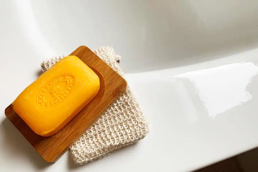 Ein Stück Seife auf einer Holzseifenablage neben einem Sisalschwamm auf dem Rand einer Badewanne.
