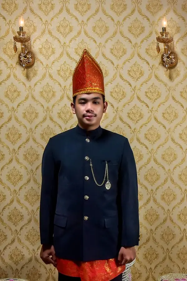 Pria muda mengenakan pakaian adat, berdiri di depan dinding bermotif.
