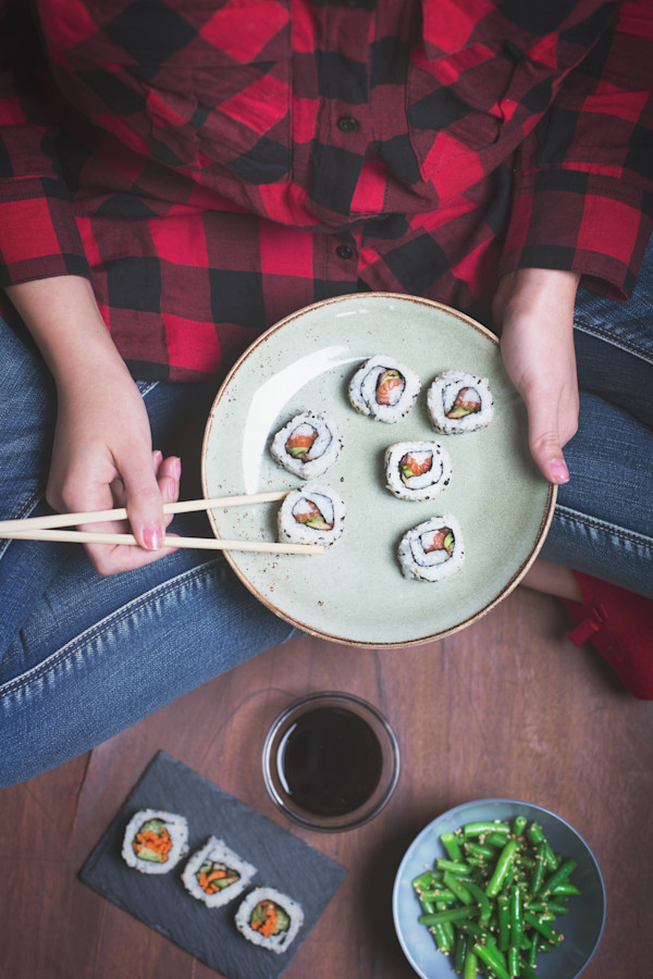 Person in a red and black plaid shirt holding chopsticks with sushi on a plate, more sushi on a slate, and a bowl of green beans on a wooden surface.