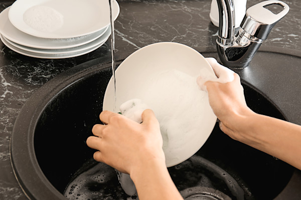 A person washing a white bowl in a black kitchen sink with soapy water, with stacked plates nearby.