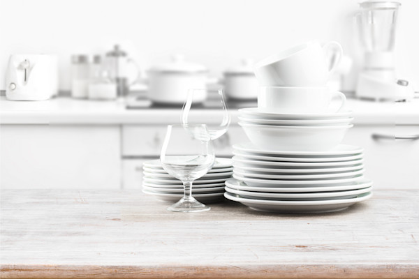 A stack of clean white dishes and cups on a kitchen counter with kitchen appliances in the background.