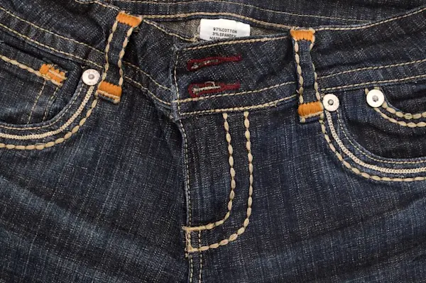 Bagian depan celana jeans dengan kancing dan jahitan.