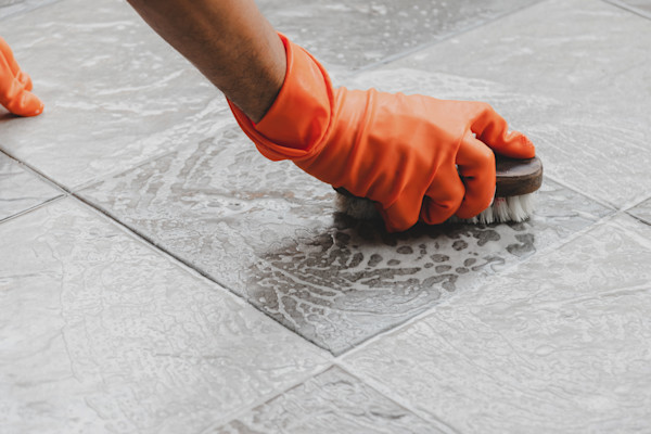 A person wearing orange rubber gloves scrubbing a tiled floor with a brush.