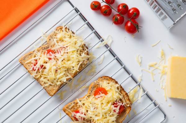 Zwei Scheiben Toast mit Tomaten und geriebenem Käse auf einem Backrost, Käseraspel daneben, ganzer Käseblock und Strauchtomaten rechts, Reibe links im Bild.