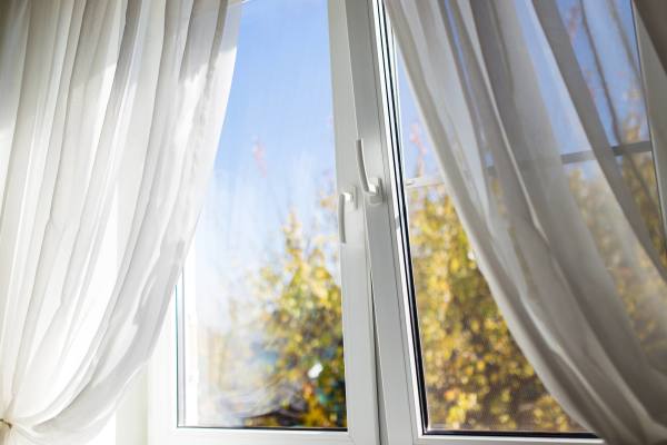 Ein sauberes, geöffnetes Fenster mit weißen Vorhängen und Blick auf Bäume im Sonnenlicht.