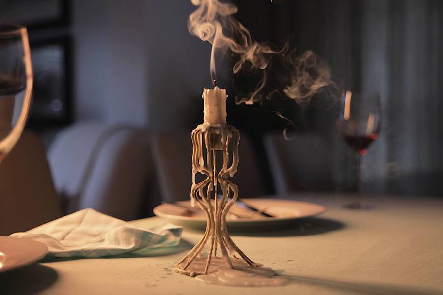 A candle with melted wax on a dinner table with smoke rising from it, a wine glass, and a napkin in the background.