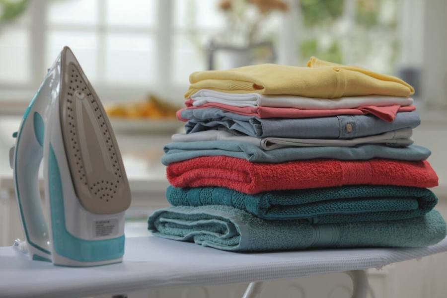 A steam iron next to a neatly stacked pile of ironed clothes and towels on an ironing board.