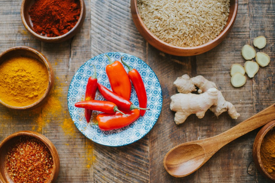 A variety of spices and rice on a wooden table, including red chilli peppers on a decorative plate, ginger root, and bowls of turmeric and other ground spices.